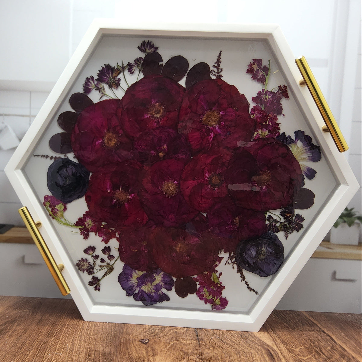 Red rose and purple ranunculus bouquet encased in clear epoxy resin inside a pressed flower hexagon ottoman tray with gold handles on a wood countertop