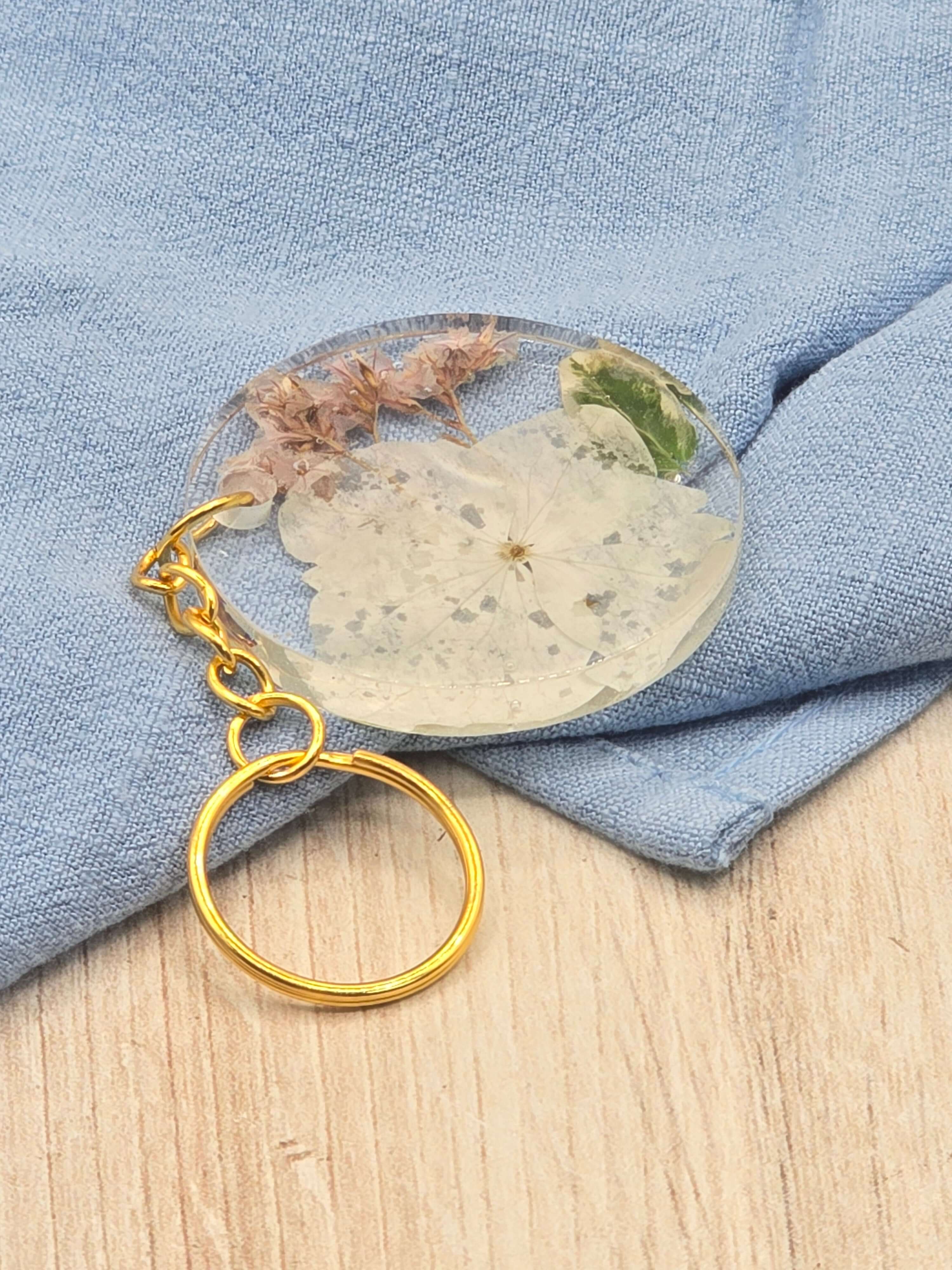 A round keychain with a gold keyring, featuring a preserved bouquet of flowers encased in clear epoxy resin, displayed on a blue fabric surface.