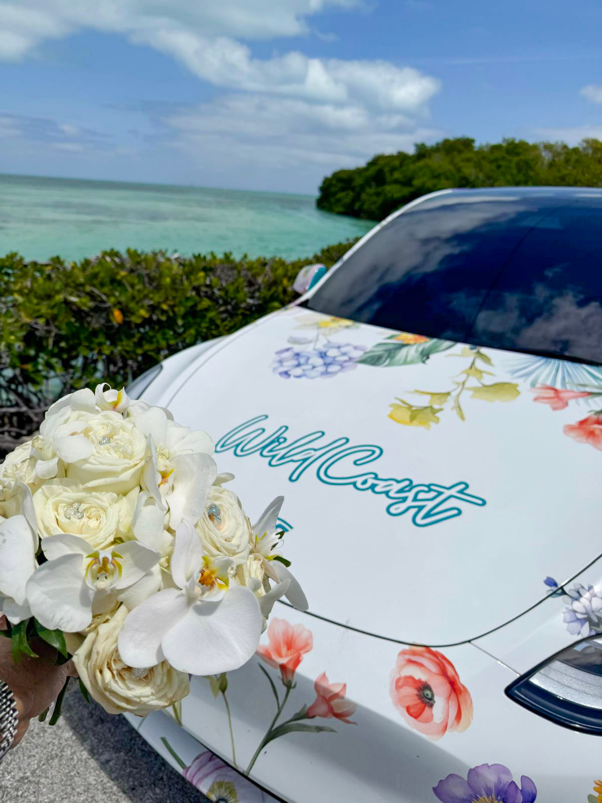 Wild Coast Flower Car with floral design and 'Wild Coast' text by a scenic waterfront with a fresh bouquet picked up from a nearby wedding.