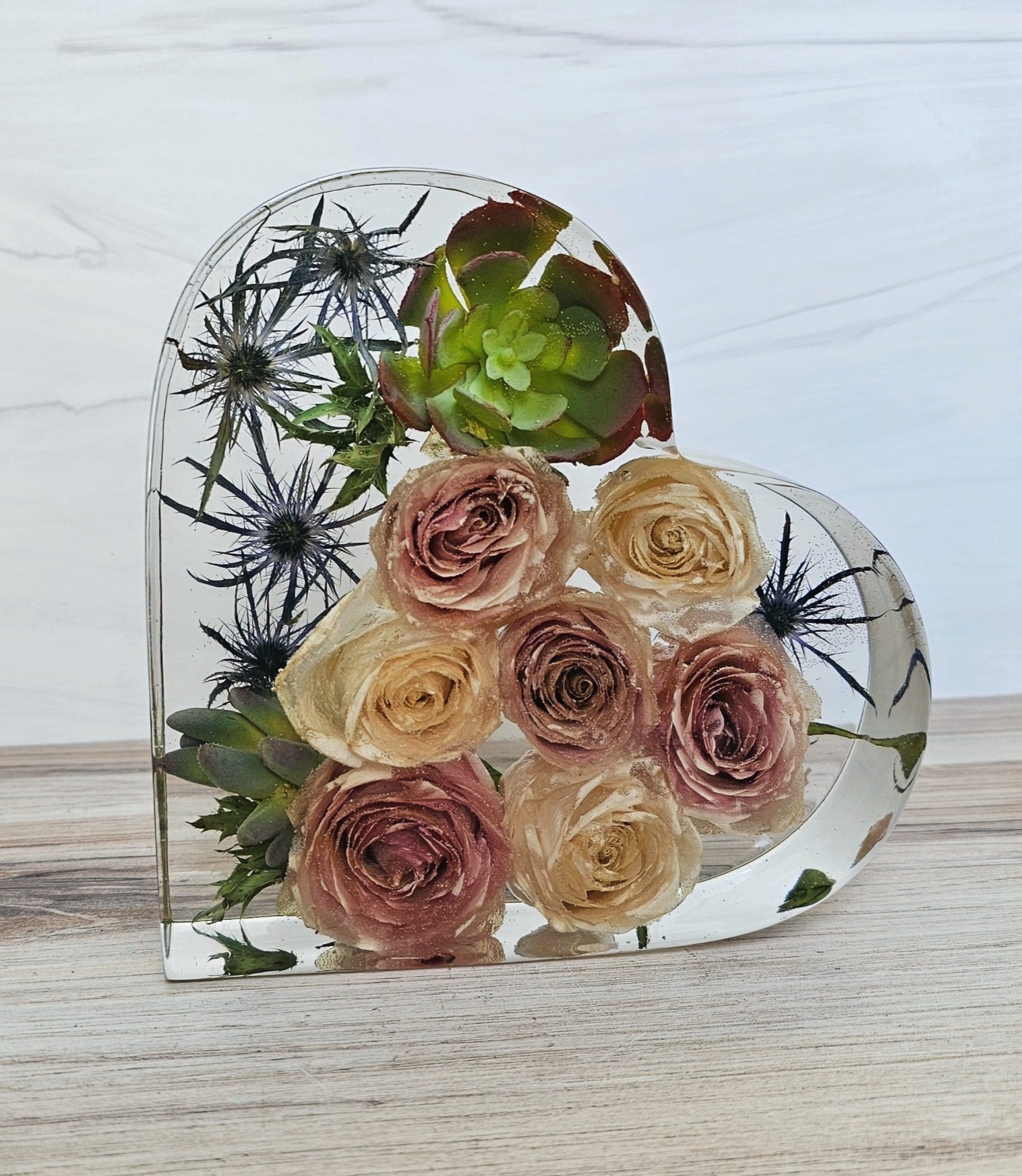 A heart-shaped resin bouquet block with preserved flowers, arranged inside with a clear resin finish, displayed on a flat surface.