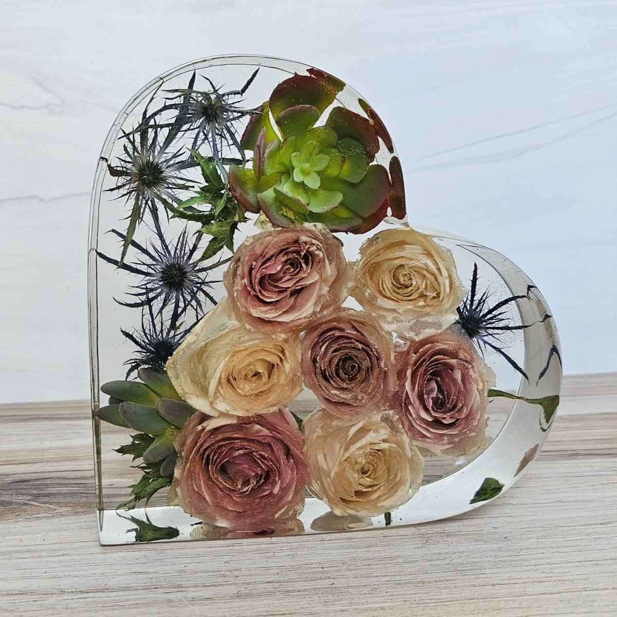 A heart-shaped resin bouquet block featuring roses, blue thistle and a fake succulent preserved inside with a clear resin finish, displayed on a flat surface.