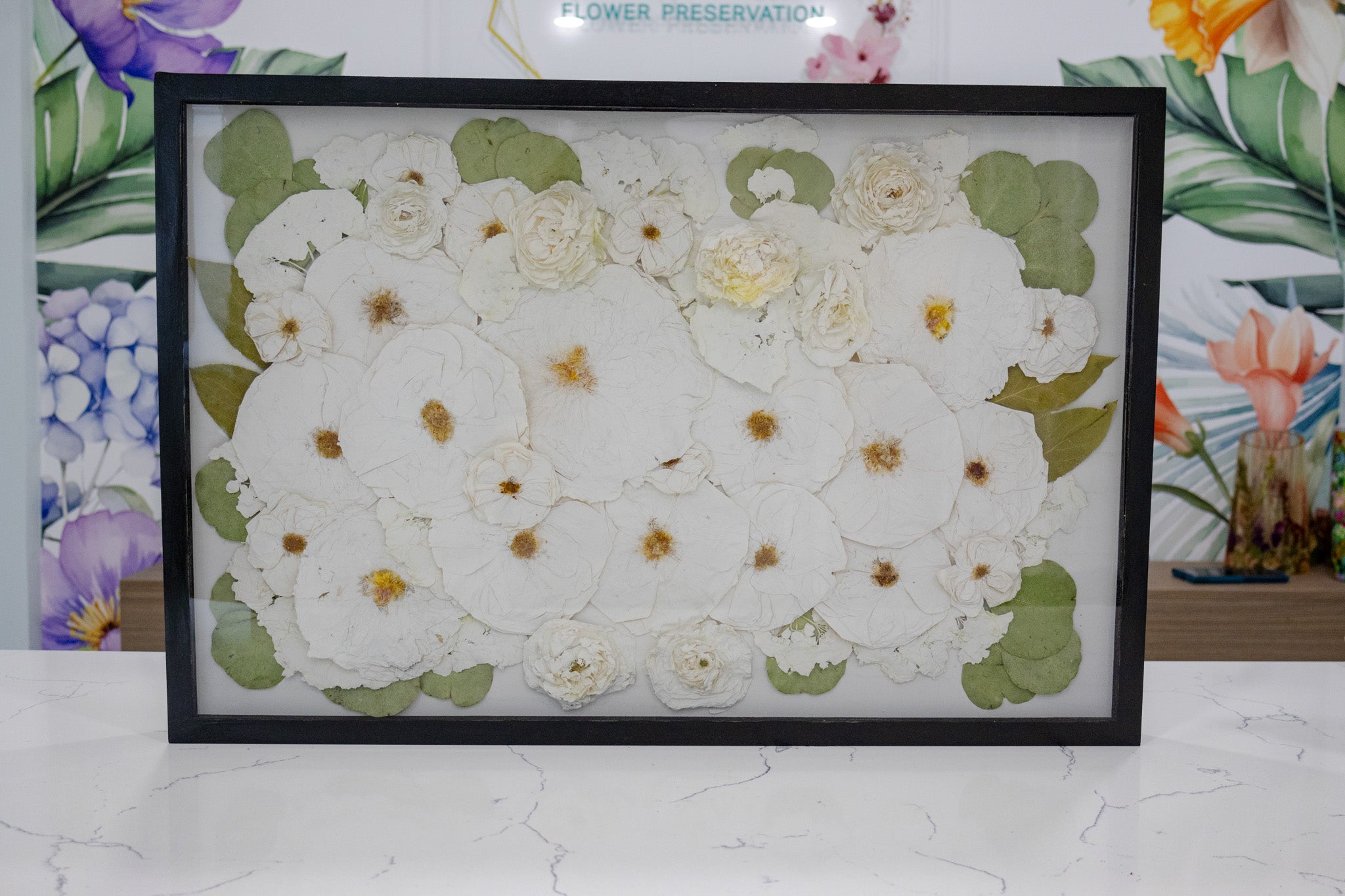A rectangular framed wood wall decor featuring a pressed flower design with a white background and green foliage accents.