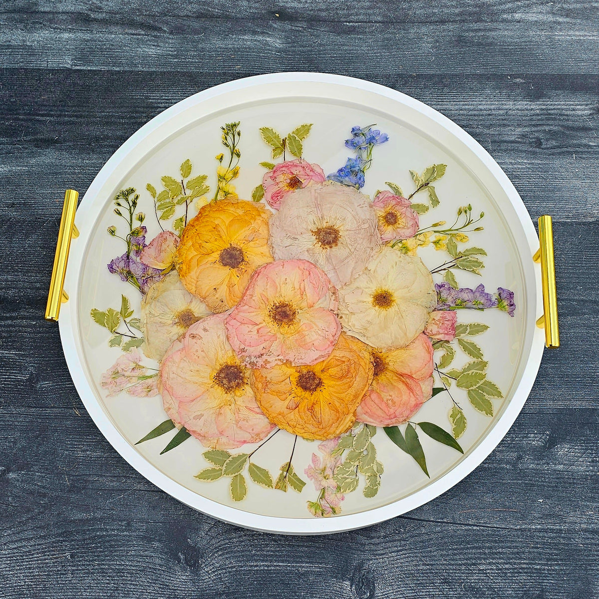 Square close-up of a pressed flower round ottoman tray showing petal depth in clear resin with white edge and gold handles on a black wood background
