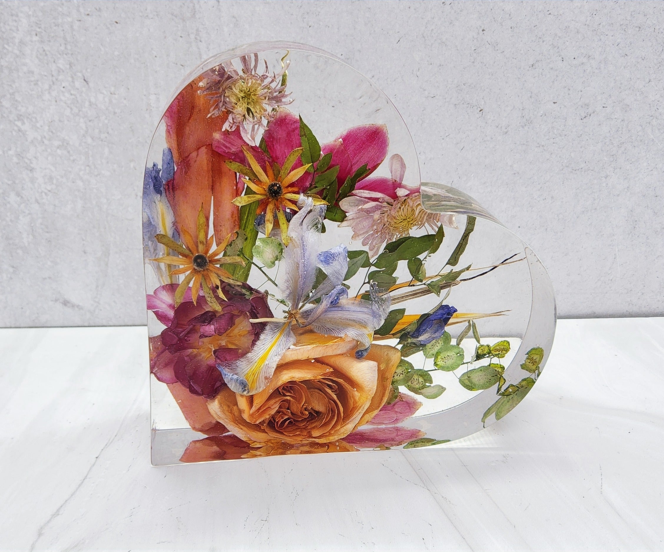 A heart-shaped resin bouquet block with preserved colorful flowers, arranged inside with a clear resin finish, displayed on a flat surface.