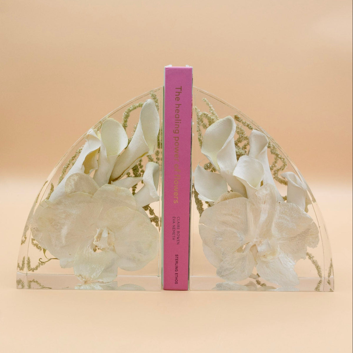 Color corrected White Calla Lillies and white orchid wedding bouquet preserved in clear resin bookends with a pink book in the middle on a tan background