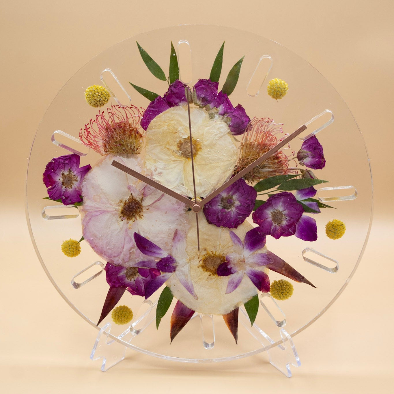 A round transparent resin wall clock with pressed flowers and green leaves in the center, displayed on a clear stand against a warm beige background.