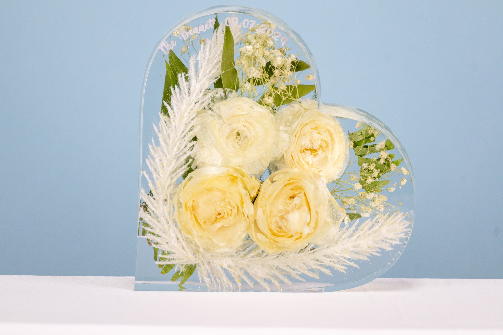 A heart-shaped resin bouquet block with preserved white roses, feathers and ranunculus flowers, arranged inside with a clear resin finish, displayed on a flat surface.