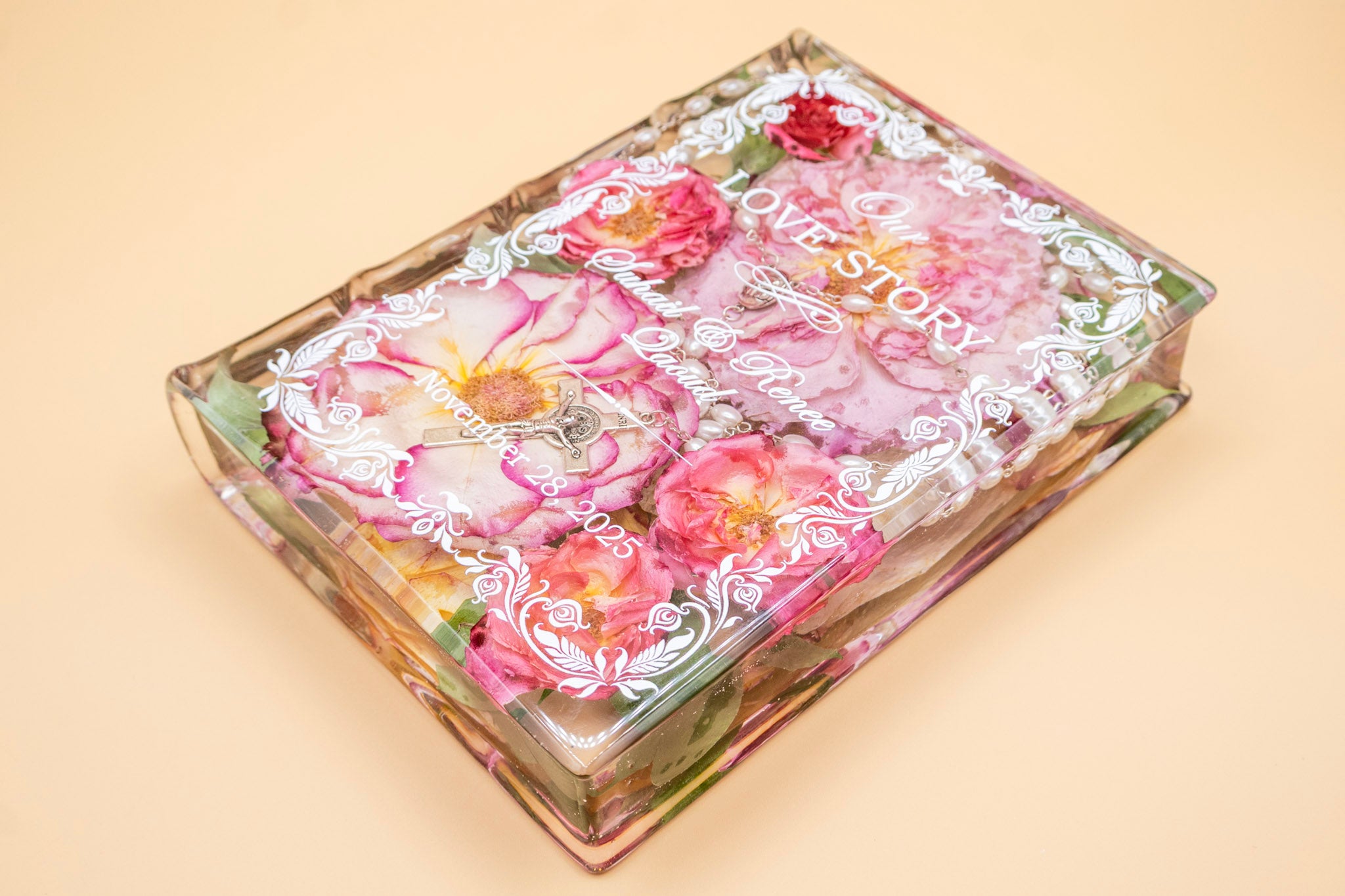 Book-shaped resin keepsake preserving pink wedding roses, pearl rosary and crucifix, with personalized names and date in white script.