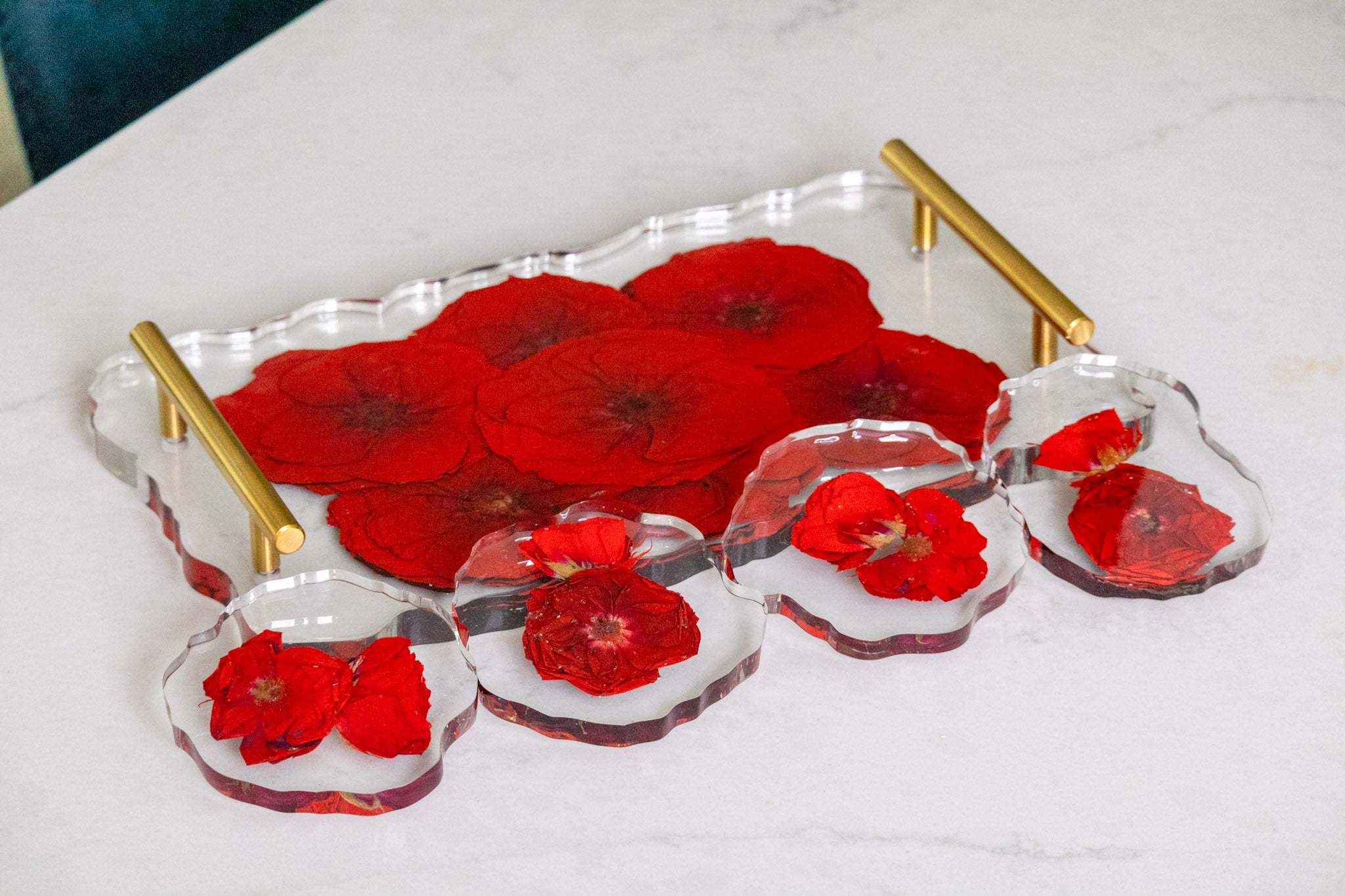 A clear acrylic serving tray set with gold handles, featuring a large rectangular tray and four matching scalloped round trays with vibrant red pressed dried flowers, set on a white marble surface.