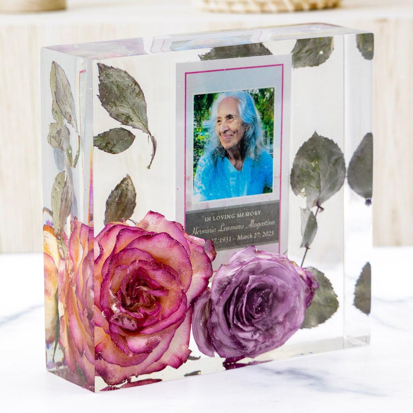 Clear resin block with preserved funeral flowers and a prayer card memorial display of a woman on a white surface