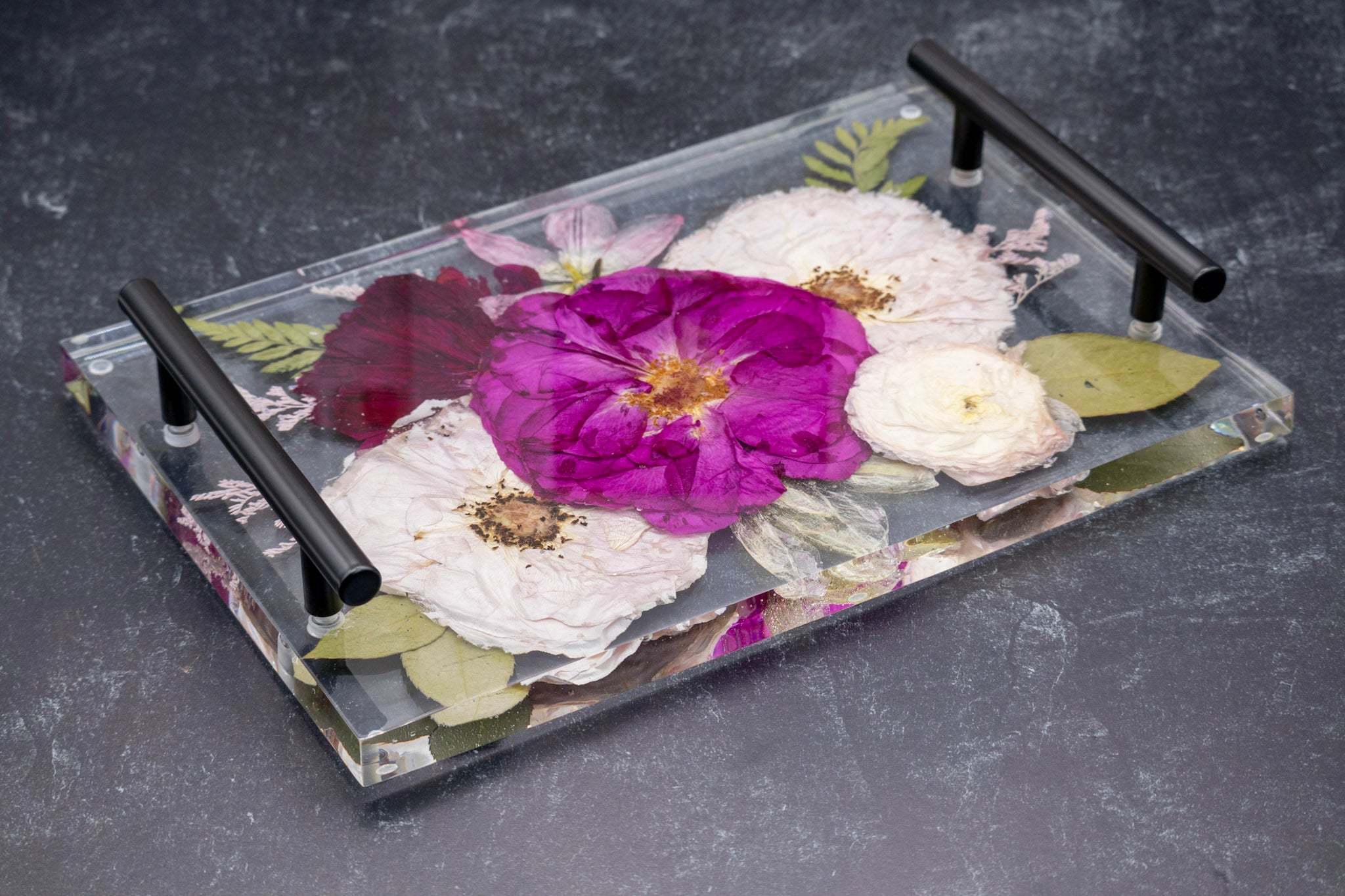 Decorative epoxy tray with pressed flower design and black handles on a dark surface