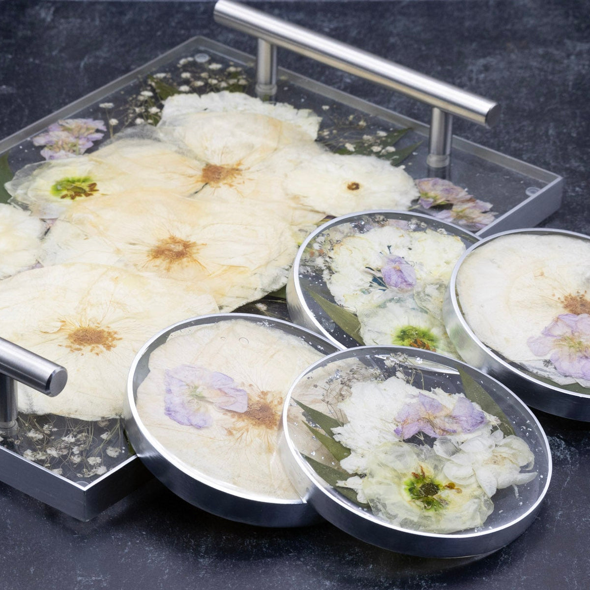 A set of clear resin keepsakes, including a rectangular tray and four round coasters with silver accents featuring dried pressed white roses and lisianthus flowers and small purple stock flowers, displayed on a dark textured surface.