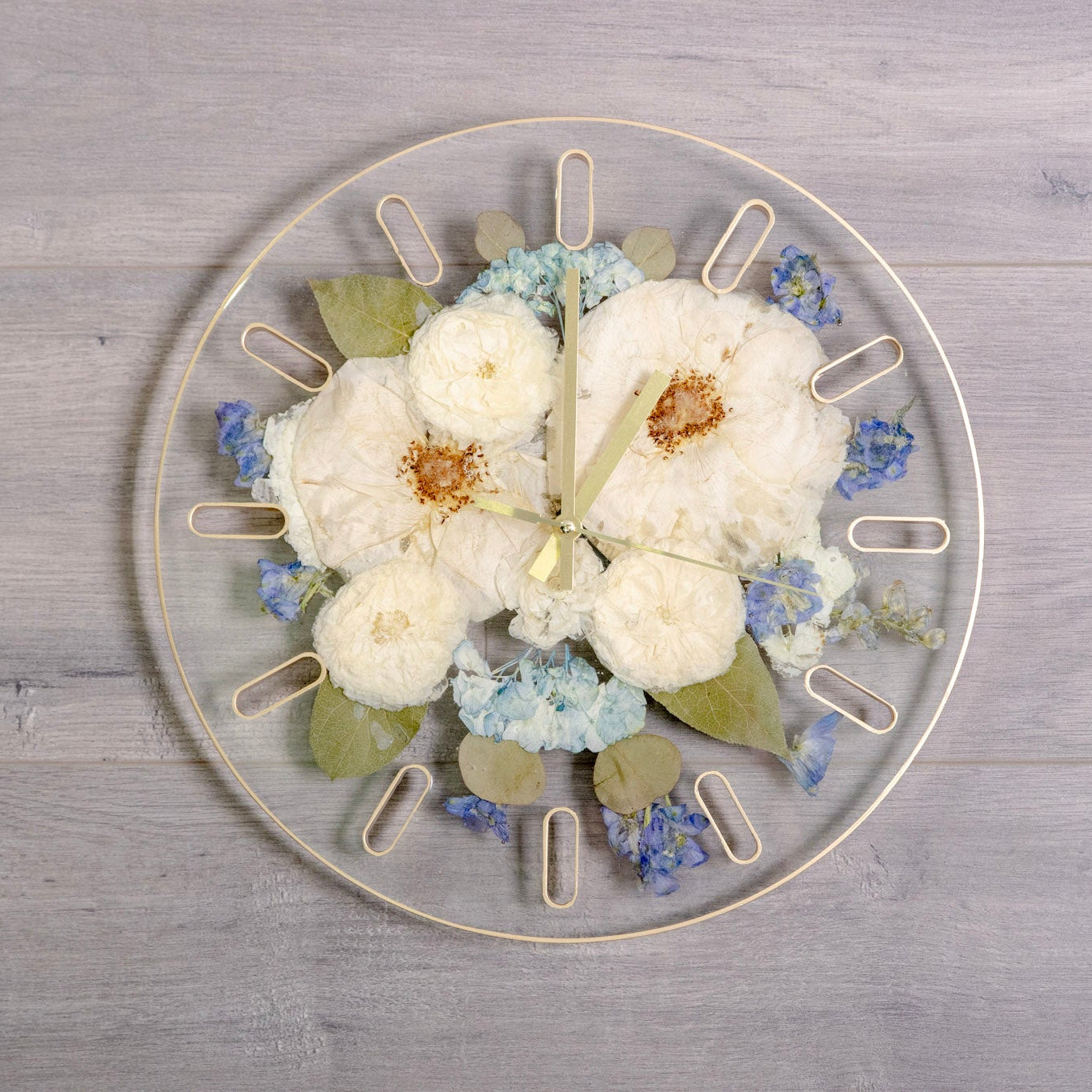 A round clear resin wall clock with pressed white rose and blue hydrangea, green leaves, and gold-tone hour markers, displayed on a light gray wood plank background.
