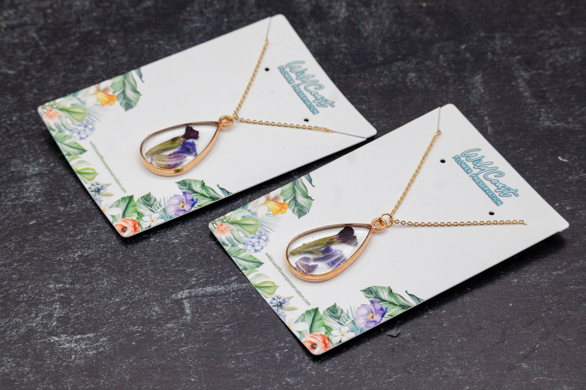 funeral flower preservation memorial necklaces Two gold necklaces with pendant flowers on floral card holders against a dark textured background.