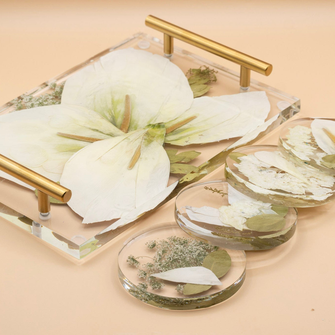 Resin flower preservation of pressed calla lily with natural color correction service encased in a serving tray with gold metal handles with 4 matching round pressed flower coaster shown on a warm beige background.