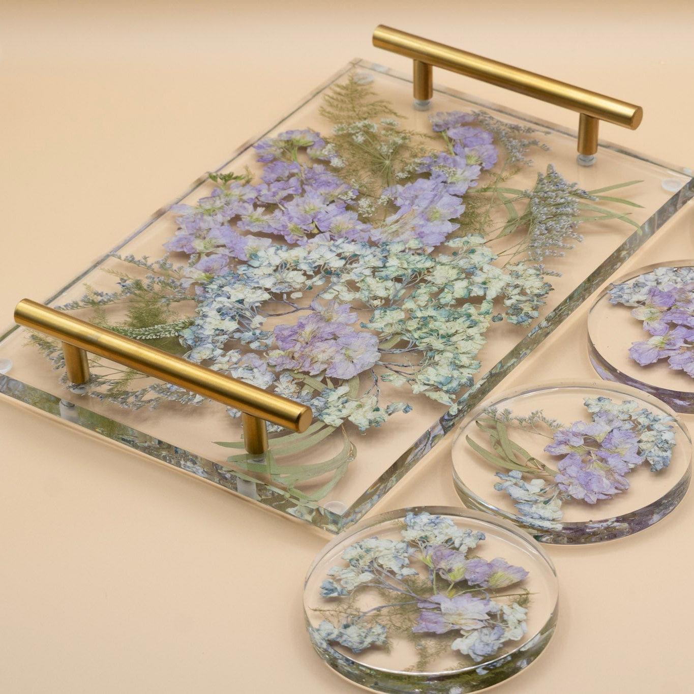 Wedding bouquet preservation of blue hydrangea and purple stock flower with pressed veronica flower and eucalyptus leaves in a clear resin serving tray with gold-tone handles, alongside four matching round coasters on a beige background.