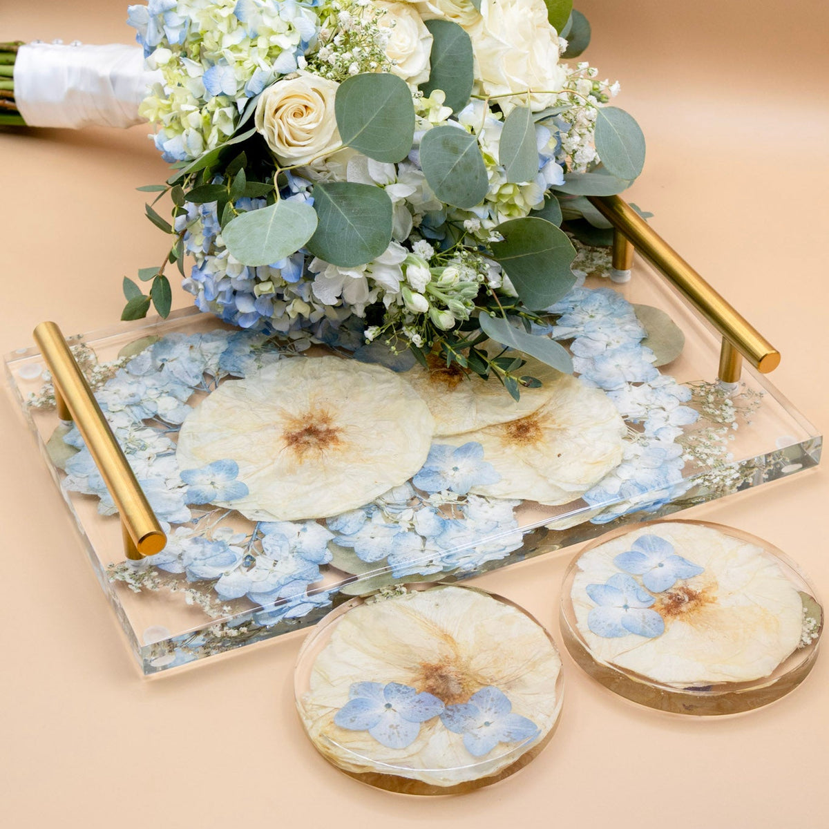 A floral arrangement with white and blue hydrangea flowers, green eucalyptus leaves and white rose with a ribbon-wrapped stem placed on a clear rectangular resin flower preservation tray filled with dried pressed flowers and gold-toned handles. Two matching round resin coasters with floral designs sit in front on a warm beige background.