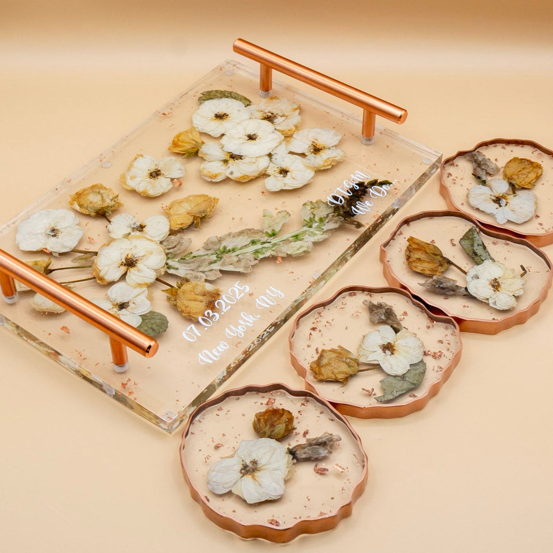 A clear resin flower preservation decorative tray with copper-toned handles and four matching rose gold geode-edge coasters, both decorated with pressed white and yellow rose flowers and green leaves, arranged on a warm beige background.