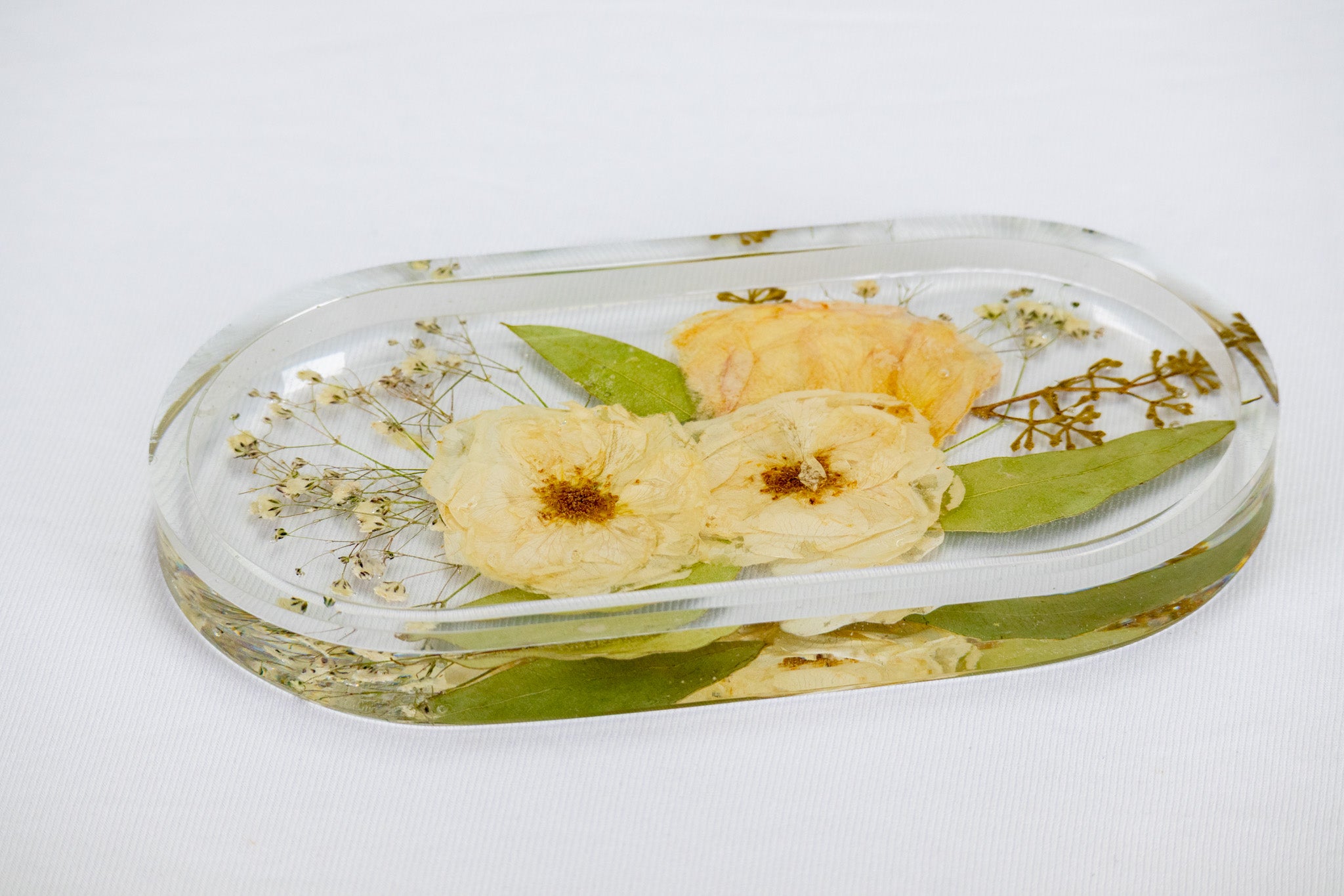 An oval-shaped trinket tray with preserved flowers, showcased on a flat surface alongside some jewelry items.