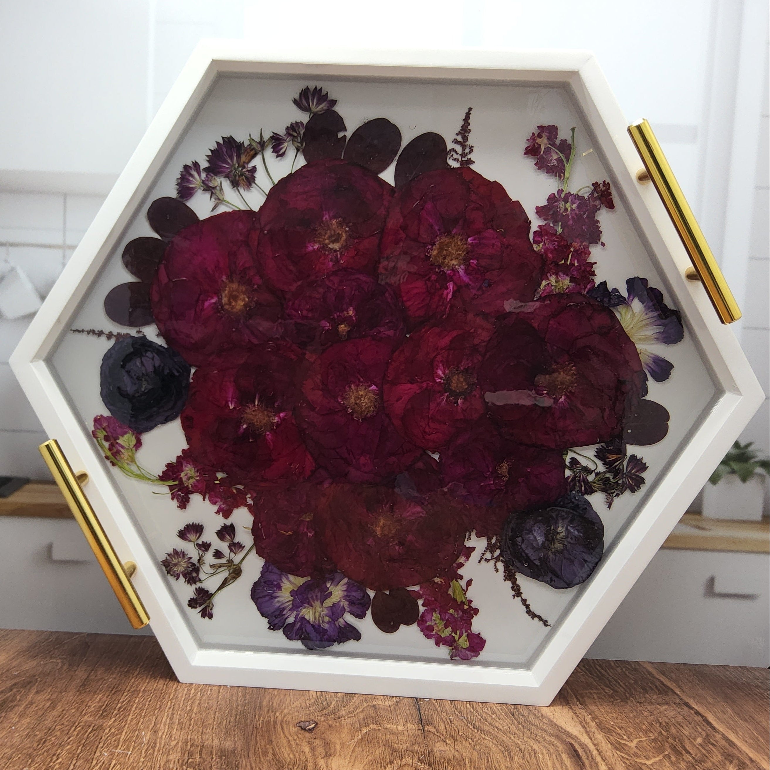 Red rose and purple ranunculus bouquet encased in clear epoxy resin inside a pressed flower hexagon ottoman tray with gold handles on a wood countertop