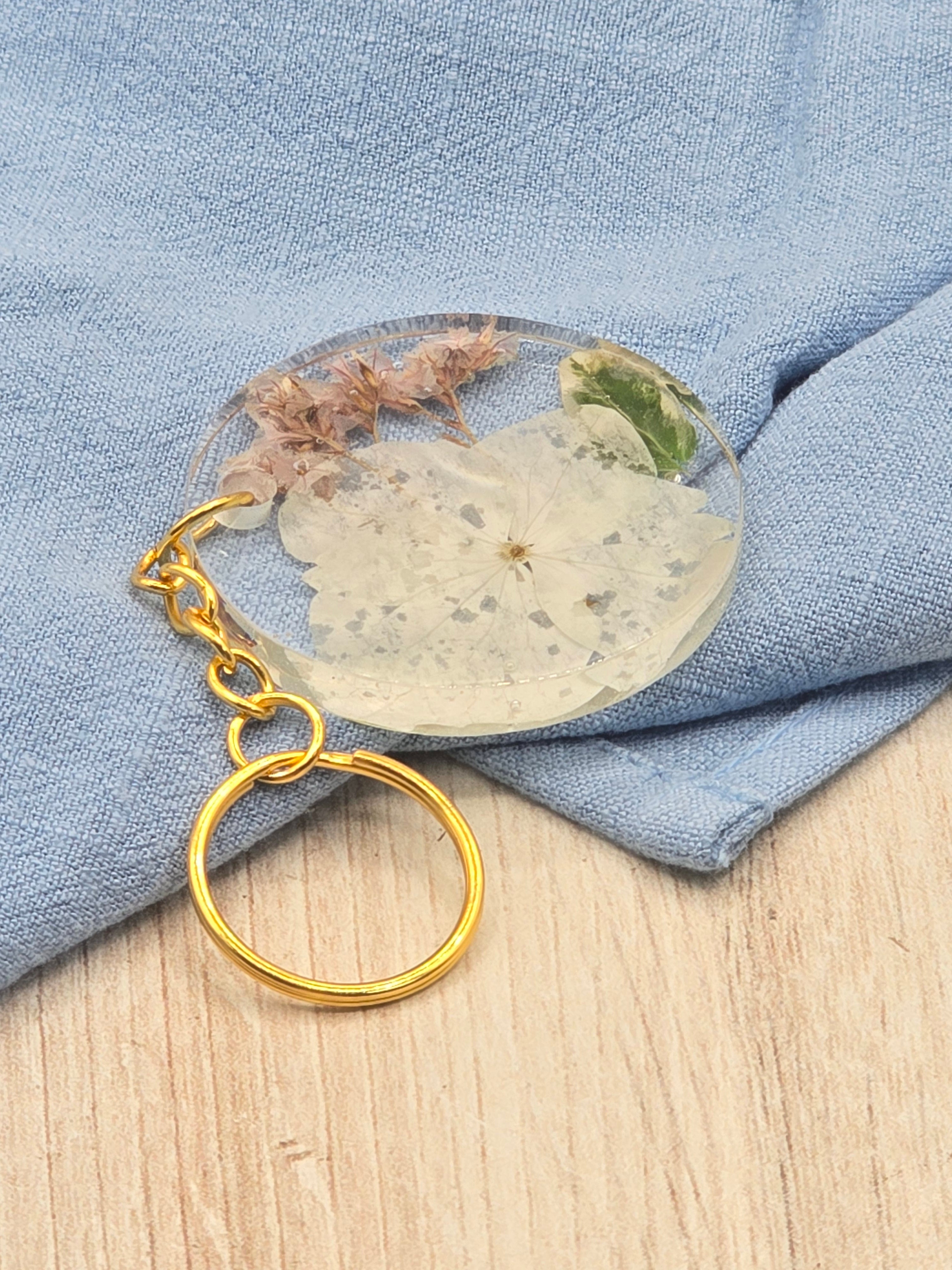 A round keychain with a gold keyring, featuring a preserved bouquet of flowers encased in clear epoxy resin, displayed on a blue fabric surface.