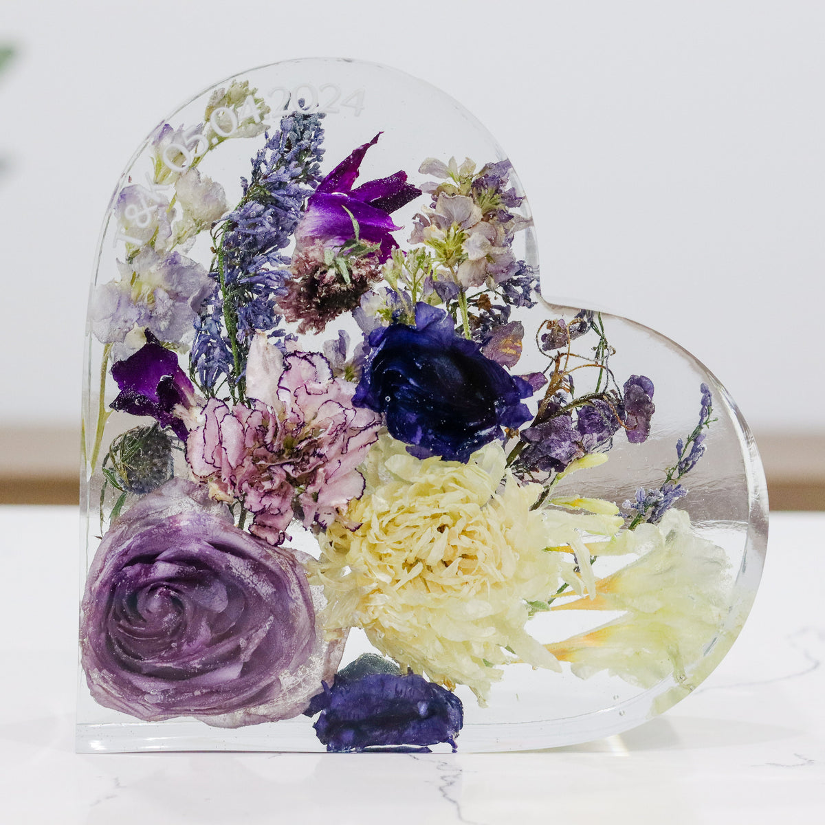 A heart-shaped resin bouquet block with preserved purple wedding bouquet flowers, arranged inside with a clear resin finish, displayed on a flat surface.
