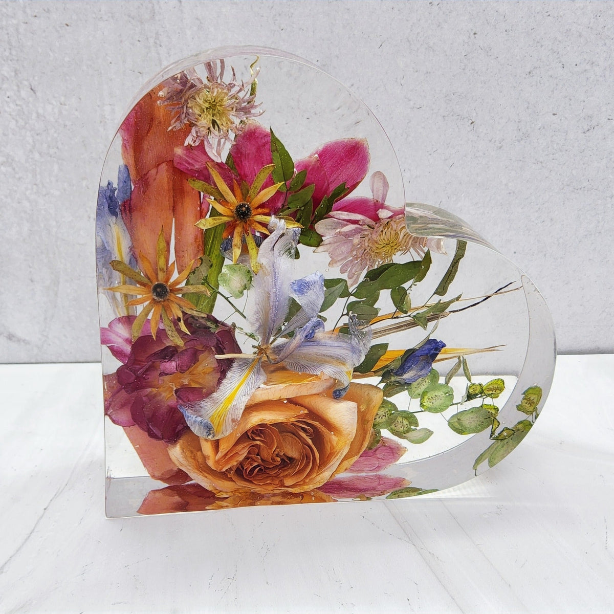 A heart-shaped resin bouquet block with preserved colorful flowers, arranged inside with a clear resin finish, displayed on a flat surface.
