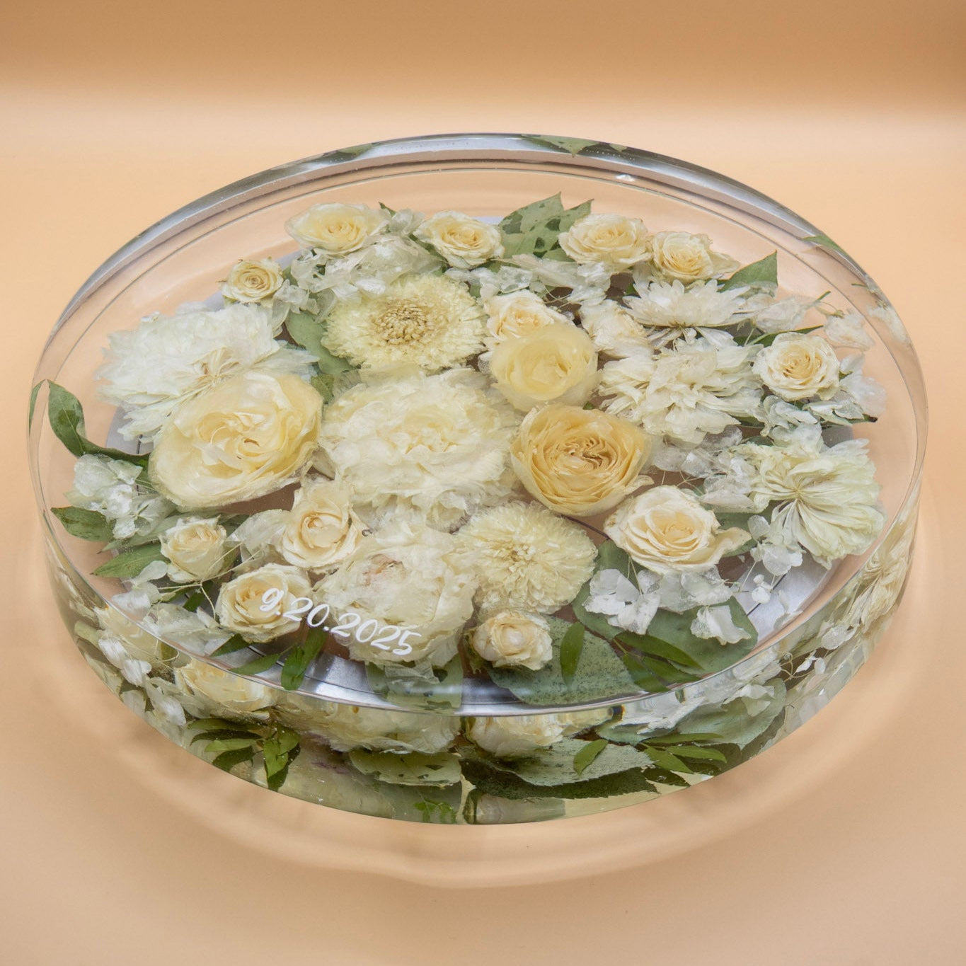 A preserved flower lazy susan whole white and pale yellow dried flowers and green leaves, encased in resin lazy susan with rotating base set against a solid beige background.