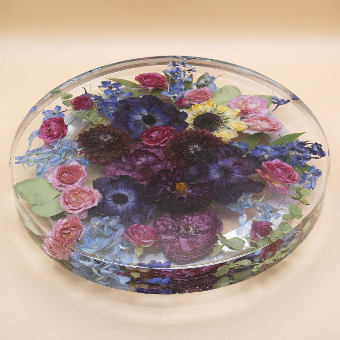 A preserved flower lazy susan  featuring whole dried flowers and green leaves of a jewel tone bouquet of purple anemone and dahlia encased in resin lazy susan with rotating base set against a solid beige background.