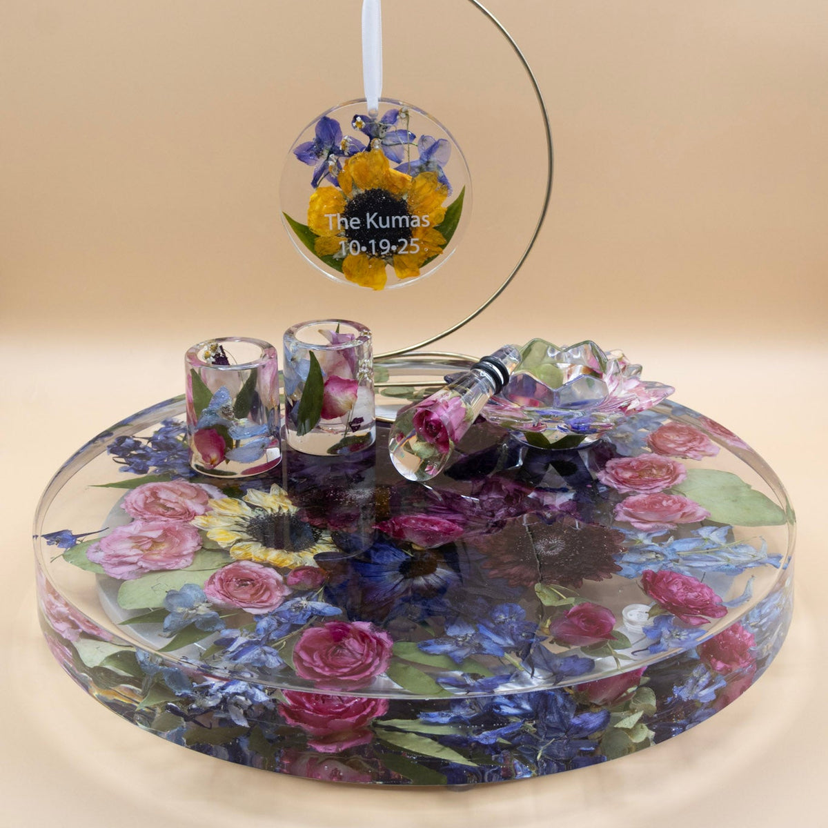 A preserved flower custom set featuring a pressed flower ornament, two shot glasses, wine stopper and jewlery cup with whole dried flowers and green leaves of a jewel tone bouquet of purple anemone and dahlia encased in resin lazy susan with rotating base set against a solid beige background.