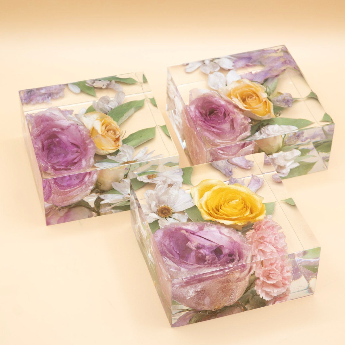 Three clear floral keepsake resin blocks with embedded dried flowers, including pink and yellow roses, displayed on a warm beige background.
