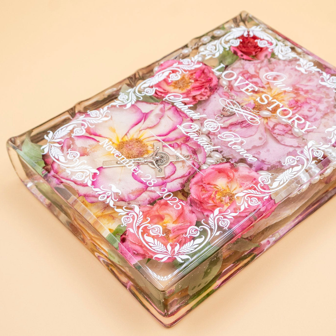 Book-shaped resin keepsake preserving pink wedding roses, pearl rosary and crucifix, with personalized names and date in white script.