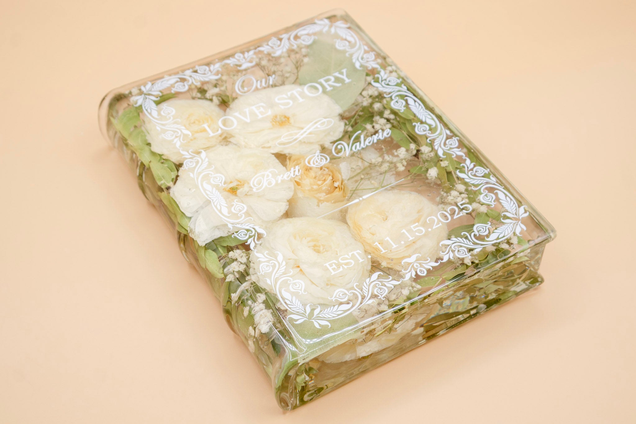 Book-shaped resin keepsake preserving white roses, baby's breath and greenery, engraved with a couple's names and wedding date.