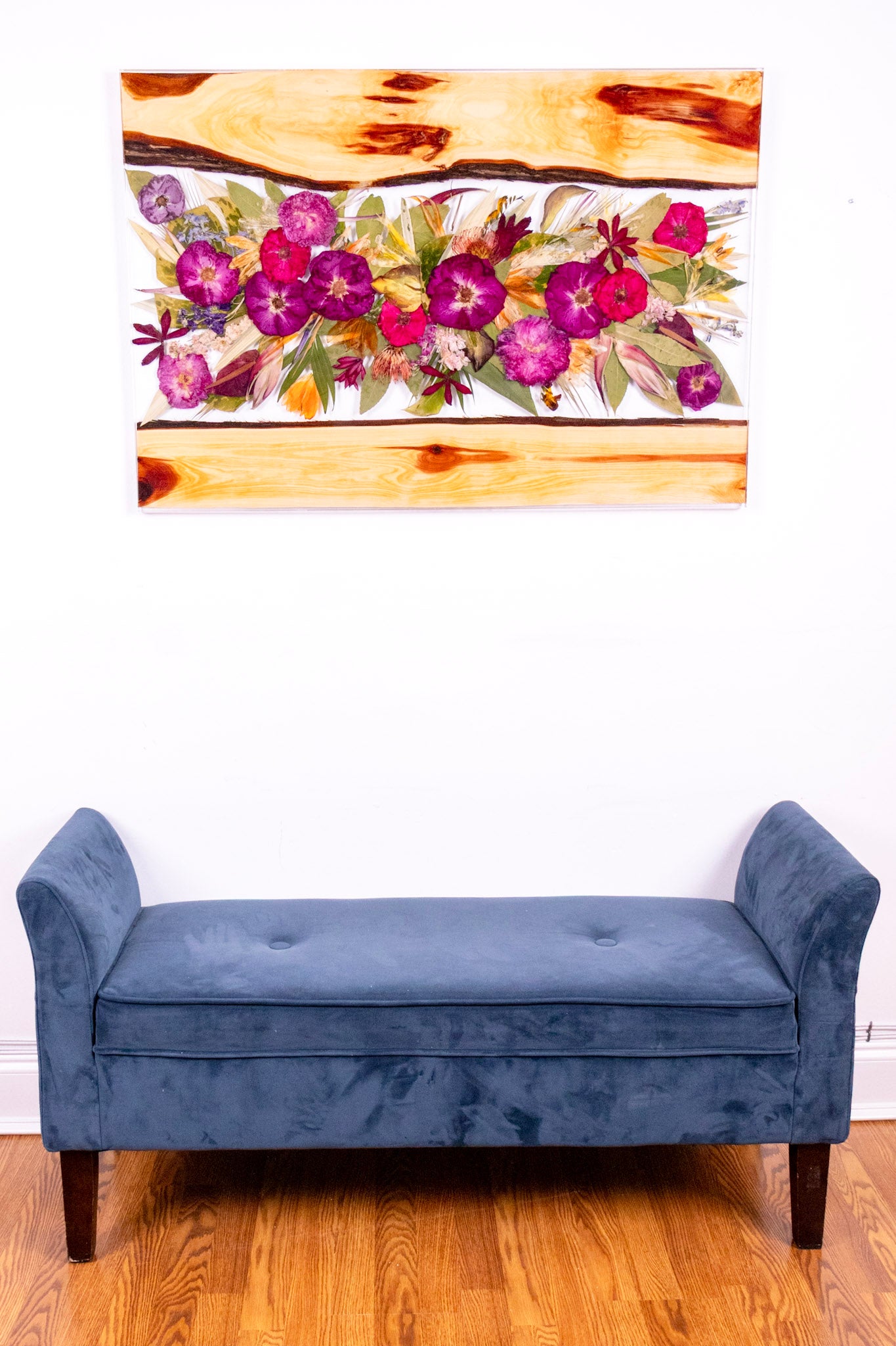 Blue bench in front of a pressed flower frame on a white wall.
