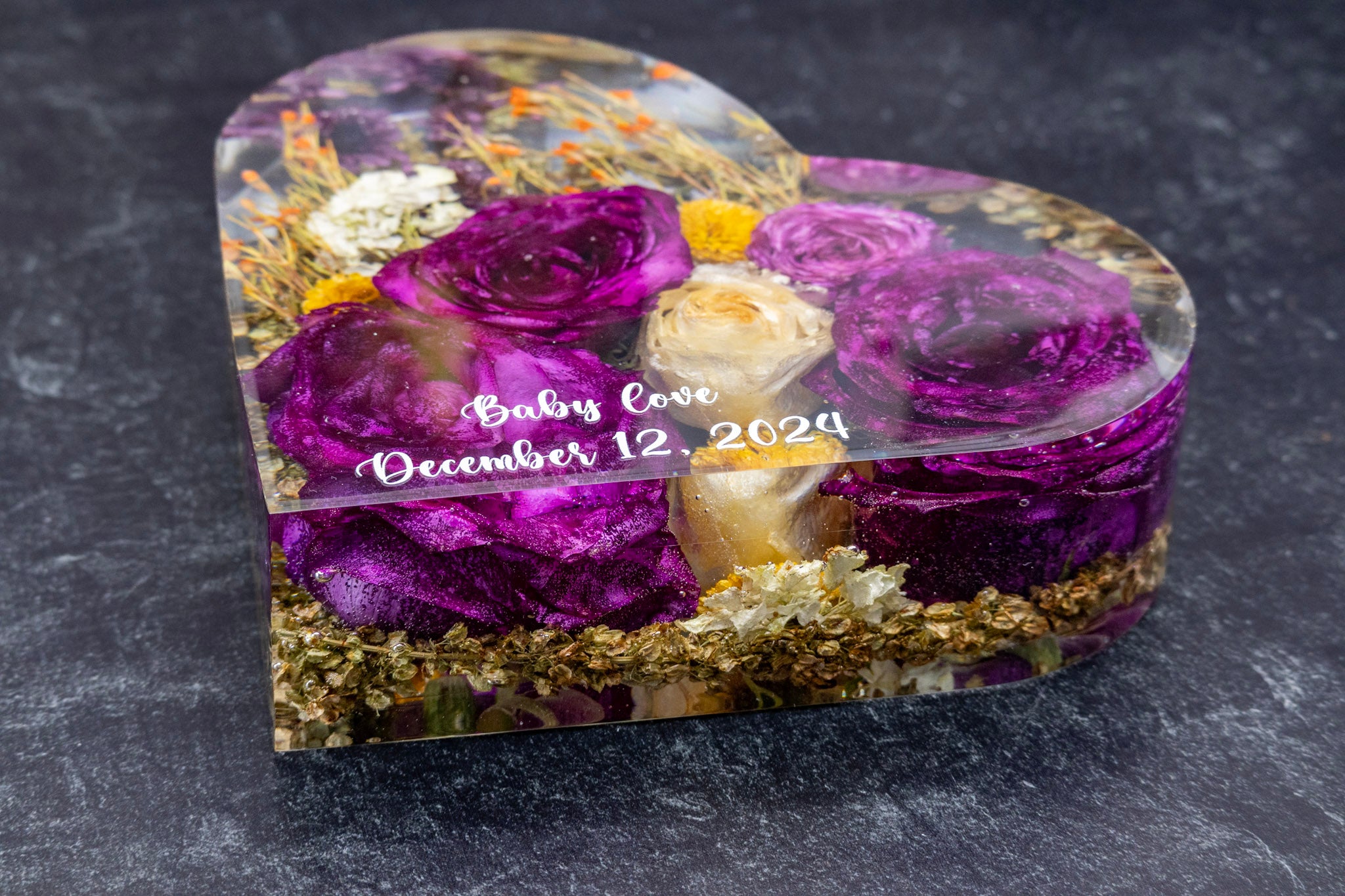 Heart-shaped resin block with text personalization saying, "baby love December 12, 2024" resin encased purple and yellow flowers on a dark background