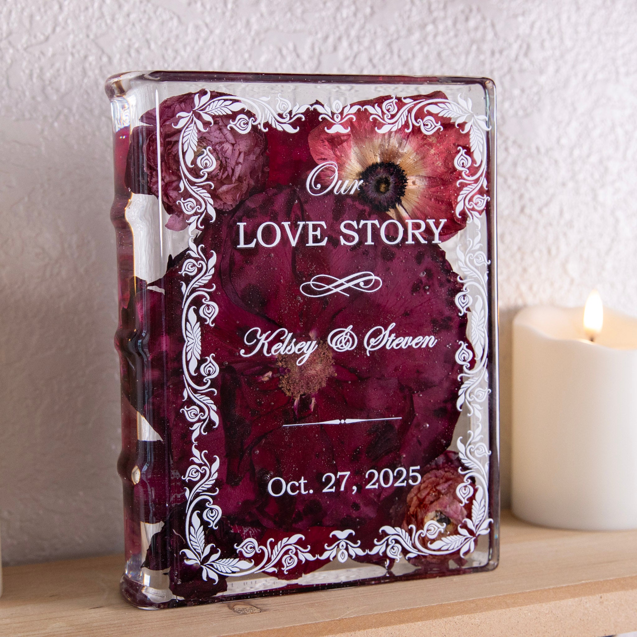Book-shaped resin keepsake with preserved burgundy wedding flowers, personalized names and date, beside a lit candle on a wooden shelf.