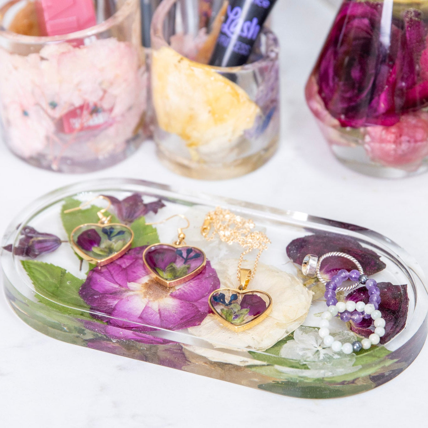 Oval resin ring dish with preserved purple and white flowers, holding heart jewelry, beside matching flower-filled resin tumblers.