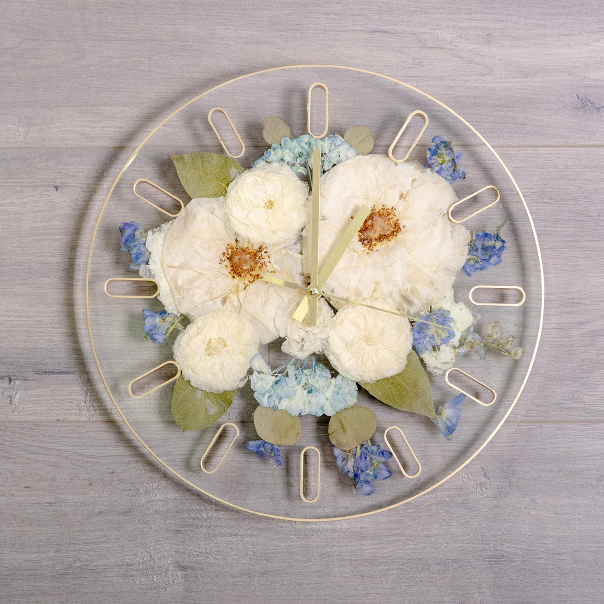 A round clear resin wall clock with pressed white rose and blue hydrangea, green leaves, and gold-tone hour markers, displayed on a light gray wood plank background.