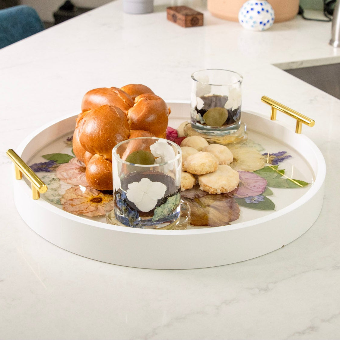 White round wood tray with gold handles and pressed flowers in clear resin with 2 cups and bead atop on a white kitchen counter.