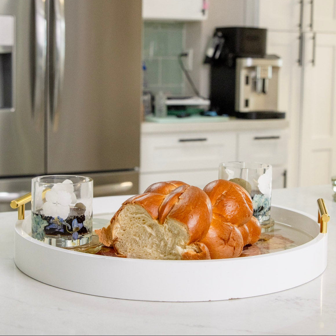 Side view of White round wood tray with gold handles and pressed flowers in clear resin with 2 cups and bead atop on a white kitchen counter.