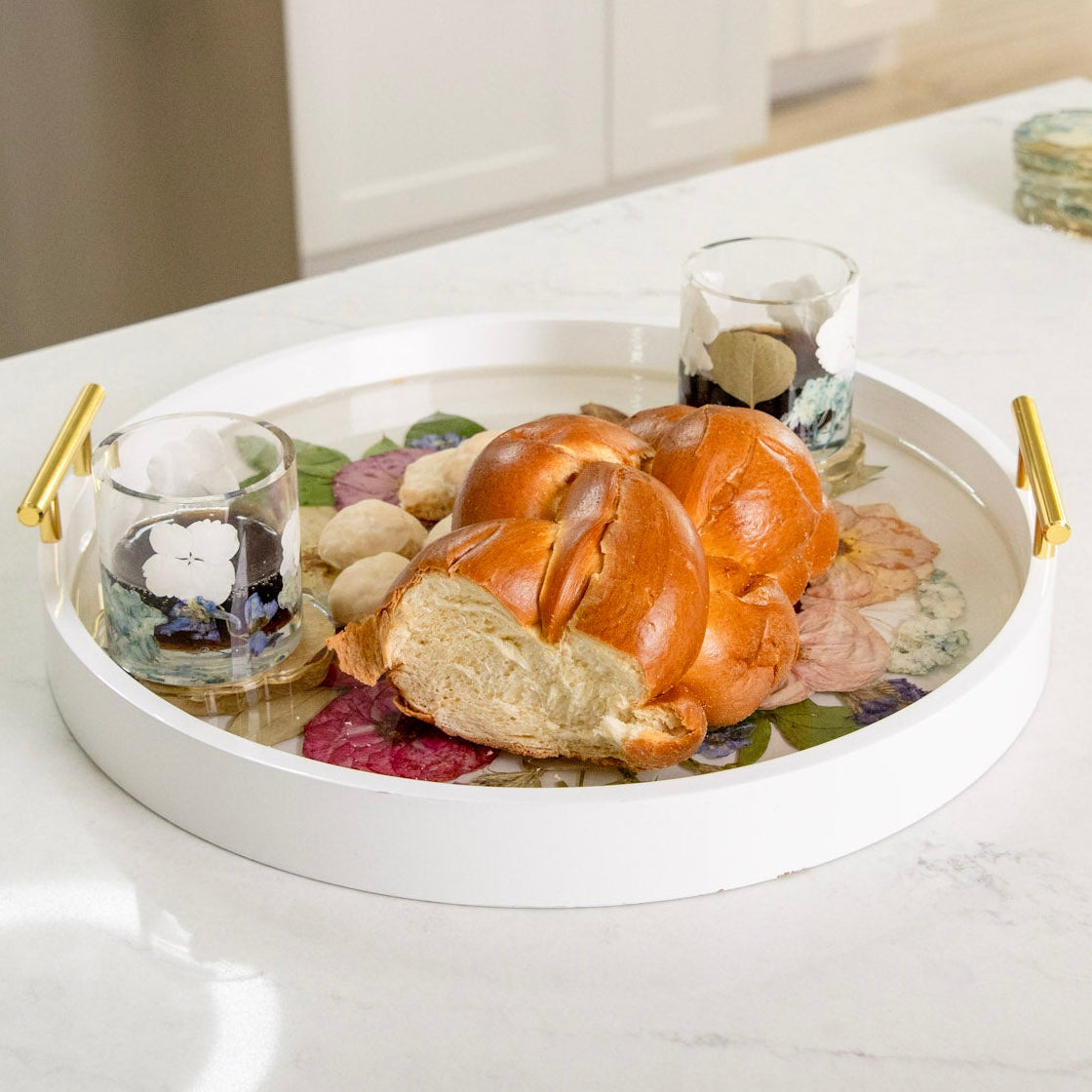 White round wood tray with gold handles and pressed flowers in clear resin with 2 cups and challah bead atop on a white kitchen counter.