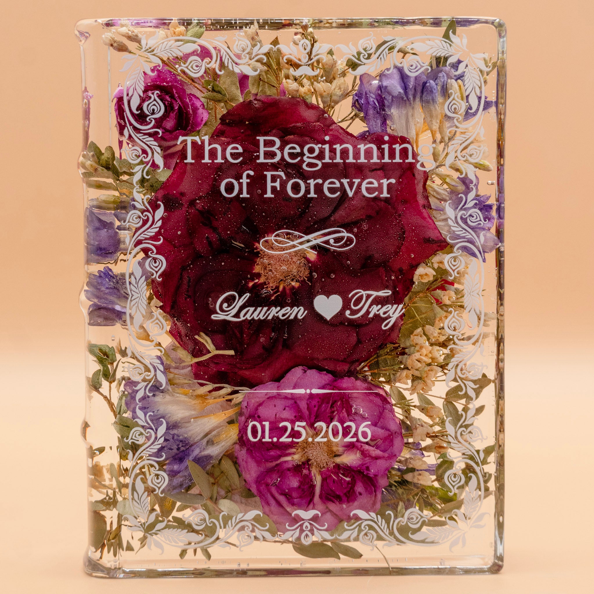 Book-shaped resin keepsake preserving burgundy and pink roses with purple blooms, engraved with a couple's names and wedding date.