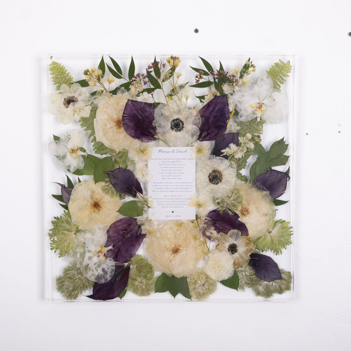 A large clear square of resin framed pressed flower arrangement on a white wall, featuring white rose, white and black anemone and deep purple calla lily with green leaves and a central printed card.