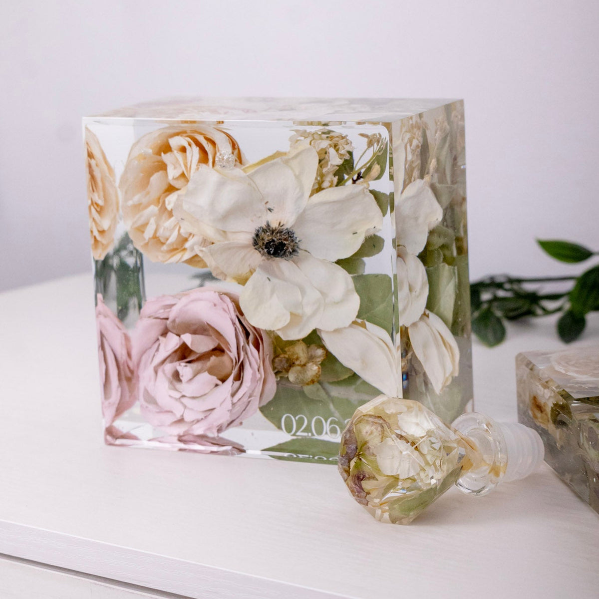 Bouquet preservation with color corrected flowers in a 5x5 resin block on a white table next to a preserved flower wine stopper