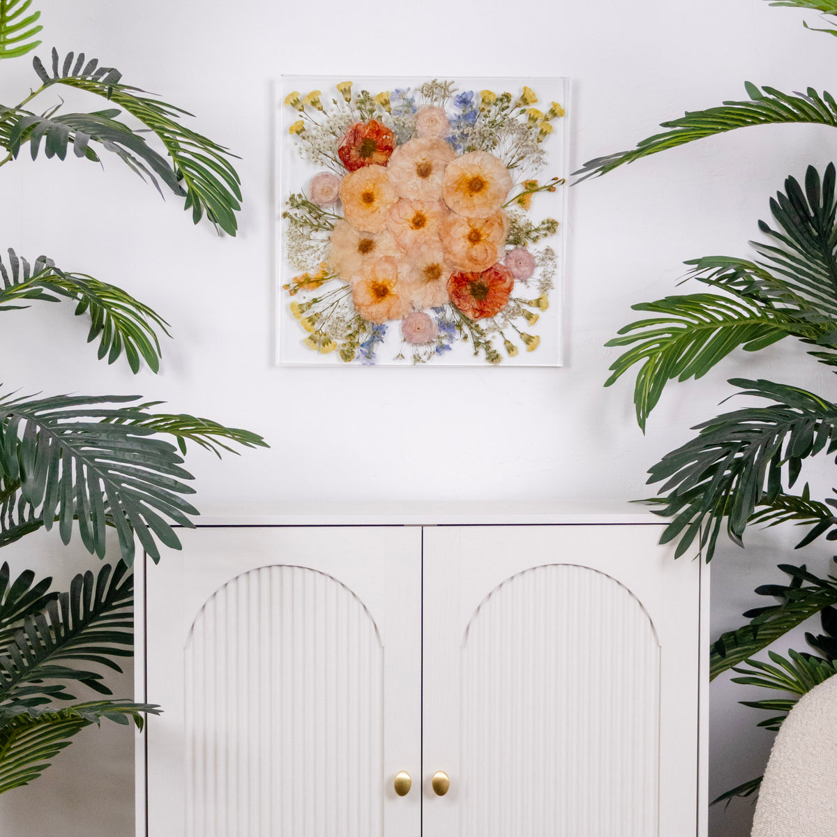 Large resin square bouquet preservation tile on crisp white wall over a white modern cabinet with tropical plants flanking