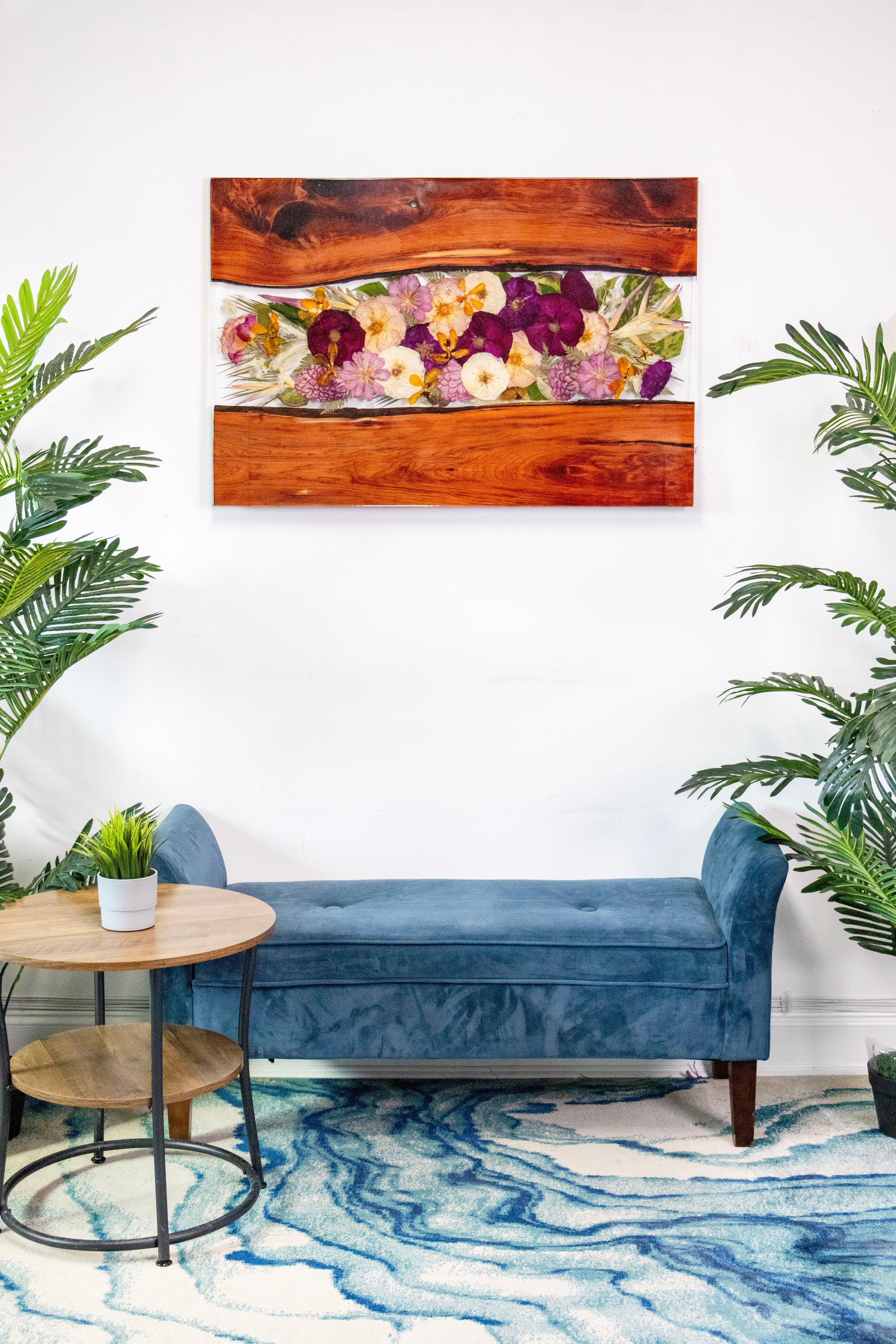 Large resin wall decor 40x27 live edge wood and pressed flower wall art hanging on the wall of a Living room with blue sofa in a room with a floral painting on the wall, plants, and a round table.