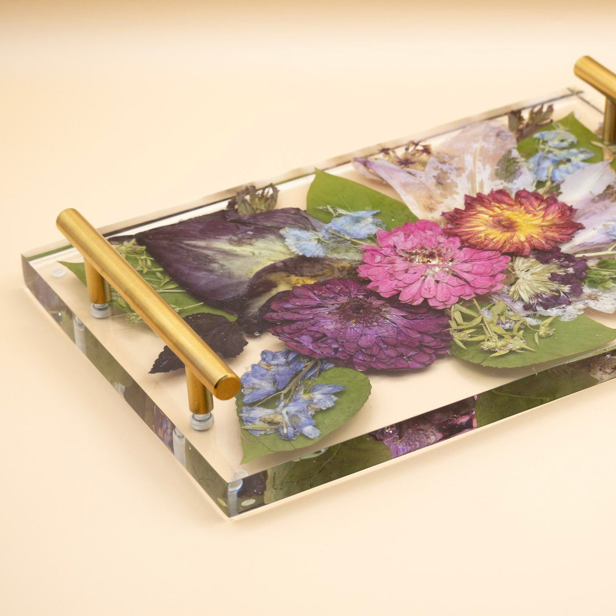 A rectangular clear resin serving tray with two gold metal handles, filled with colorful pressed flowers calla lily, zinnia and green leaves, set against a solid beige background.