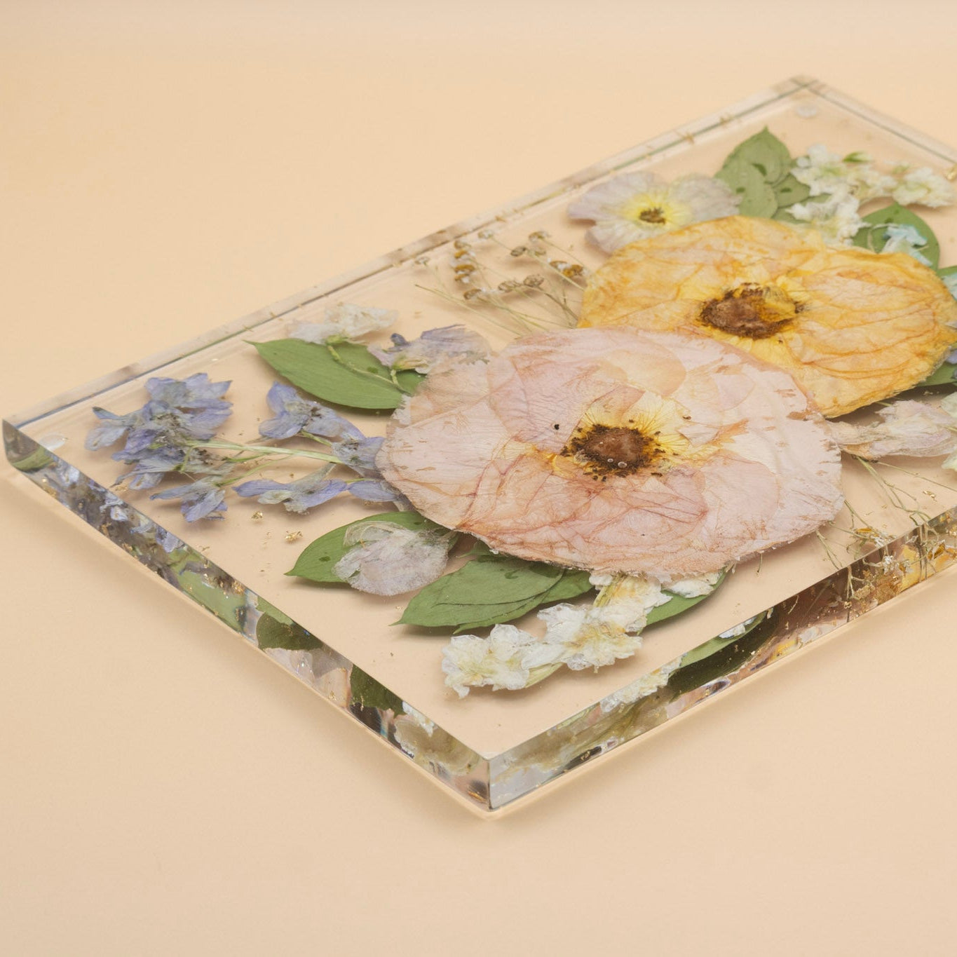 Wedding bouquet preservation rectangular clear resin tray without handles featuring pressed dried roses, including a prominent pale pink rose, a orange rose, green leaves, and small blue and white delphinium, set against a solid beige background. 