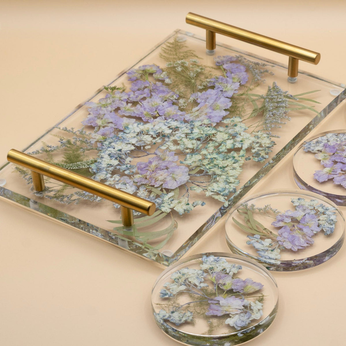 Wedding bouquet preservation of blue hydrangea and purple stock flower with pressed veronica flower and eucalyptus leaves in a clear resin serving tray with gold-tone handles, alongside four matching round coasters on a beige background.