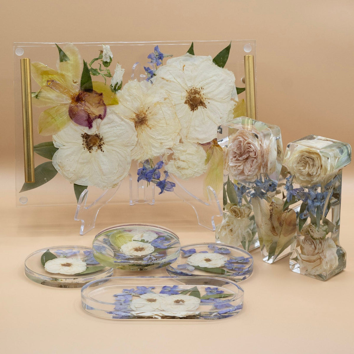 A set of resin floral preservation home decor pieces on a beige background, including a rectangular serving tray on a stand with dried white rose, letter block preservation with roses, blue delphinium and orchids, and several round pressed flower coasters and an oval ring dish.